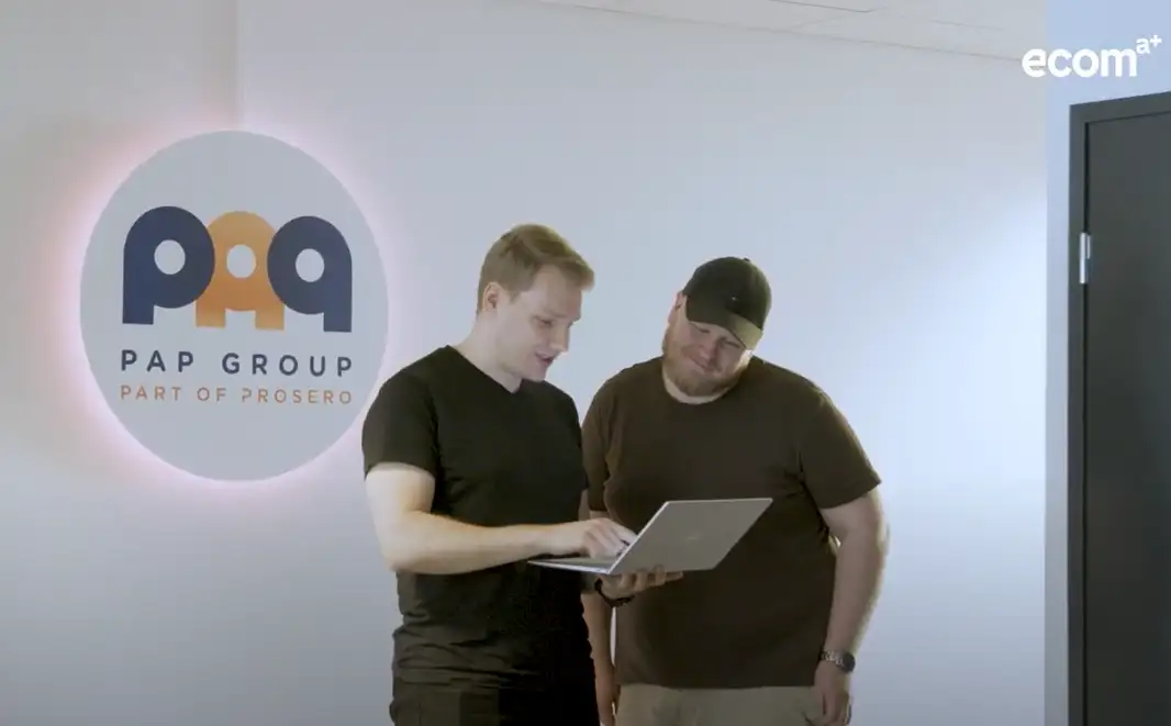 Kaksi PAP Groupin työntekijää katsoo kannettavaa tietokonetta PAP Groupin logon edessä